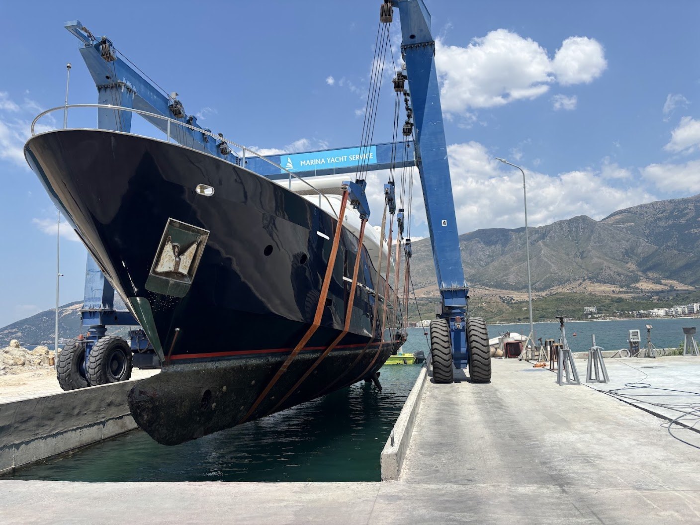 Një jaht i madh me skaf të errët i varur në rripat e travel lift-it blu me mbishkrimin Marina Yacht Service, mbi vaskën e nxjerrjes në Orikum, me kavaletë në kalatë dhe malet e Gjirit të Vlorës në sfond.